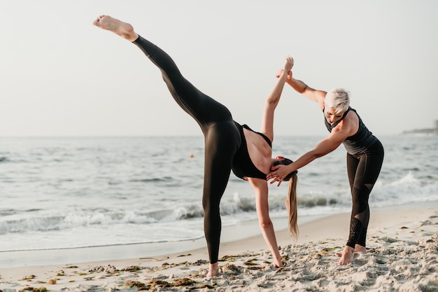 Caber mulher ajudando menina desportiva a praticar yoga asana na praia perto do oceano