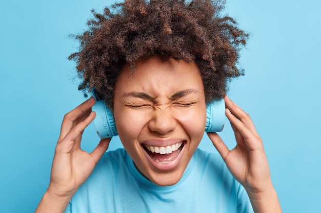 En la cabeza de una mujer afroamericana llena de alegría con el pelo rizado y tupido se ríe felizmente lleva auriculares inalámbricos y disfruta de su melodía favorita o pista de audio vestida casualmente aislada sobre una pared azul.