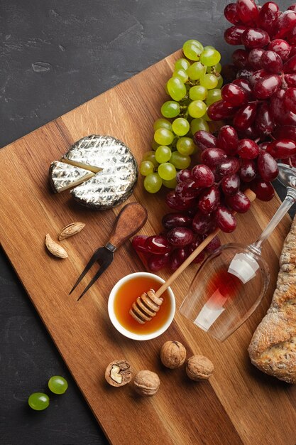 Cabeza de queso, racimo de uvas, miel, nueces y copa de vino sobre tabla de madera y fondo negro. Vista superior con espacio de copia.