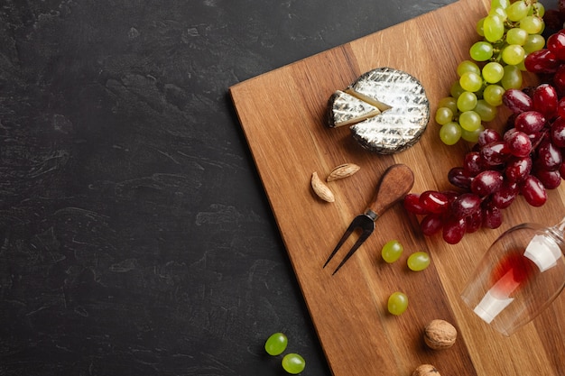 Cabeza de queso, racimo de uvas, miel, nueces y copa de vino sobre tabla de madera y fondo negro