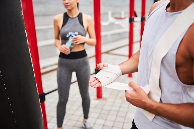 Cabeza recortada de boxeo de hombre fuerte en vendajes mientras que la mujer delgada está bebiendo agua al aire libre