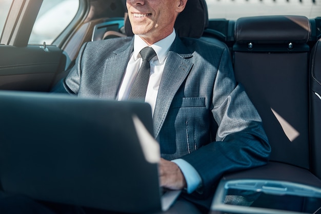 Cabeza recortada del hombre de negocios feliz que usa el cuaderno mientras es transportado por el conductor después de aterrizar en el aeropuerto