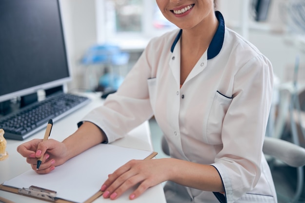 Cabeza recortada de mujer sonriente en uniforme médico haciendo notas en papel mientras está sentado en la mesa con la computadora