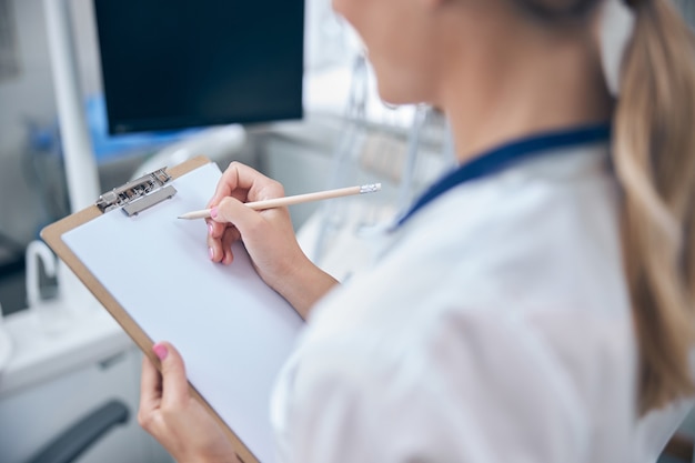 Foto cabeza recortada de mujer en uniforme médico tomando notas sobre la salud del paciente durante la jornada laboral en la clínica