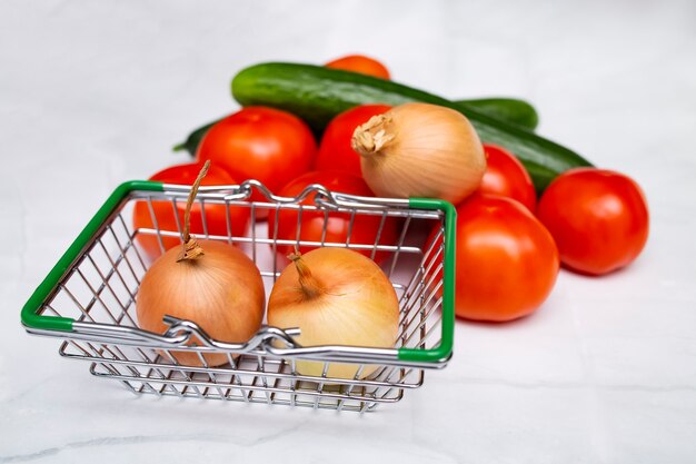 Cabezas de cebolla, tomates frescos y pepinos en canasta metálica de supermercado.