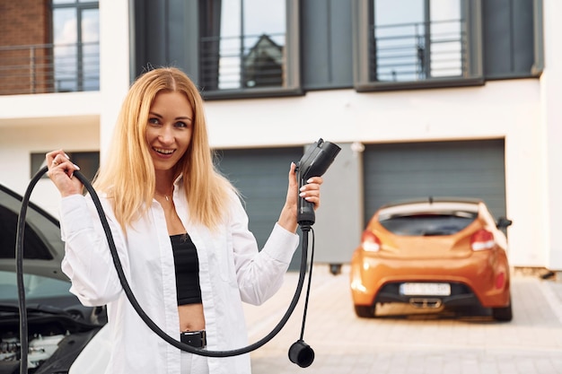 Con cable de carga en las manos Mujer joven vestida de blanco está con su coche eléctrico durante el día