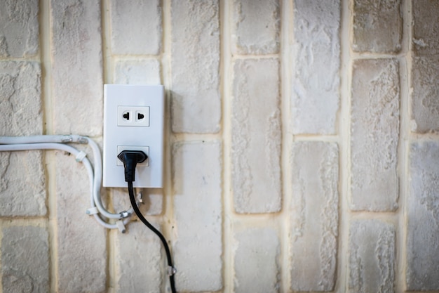 El cable negro del cable de alimentación está enchufado en el tomacorriente de pared en la pared de ladrillo de yeso blanco con espacio de copia.