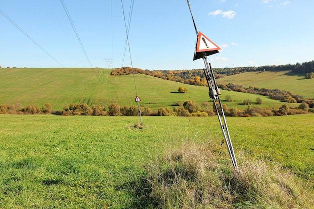 Cabos de aço no poste de eletricidade de energia com triângulos de advertência vermelhos com ponto de exclamação - perigo, alta tensão, pequenas colinas com fundo de prados verdes.