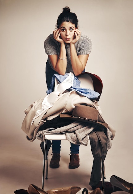 ¿Cabrán todos en mi armario? Foto de estudio de una mujer joven que elige ropa apilada en una silla contra un fondo marrón.