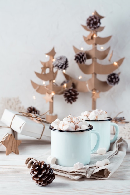 Cacau e marshmallows em canecas, presentes de artesanato de Natal e árvores de Natal em uma mesa de madeira branca. Férias ecológicas. Visão vertical