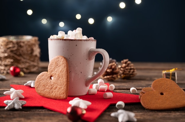 Cacau quente coberto com pequenos marshmallows em uma xícara rústica colocada sobre uma mesa de madeira cheia de decorações do feriado de Natal e grandes biscoitos em forma de coração.