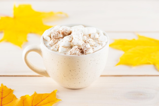 Cacau quente com marshmallows em uma caneca branca em uma mesa de madeira com folhas de outono