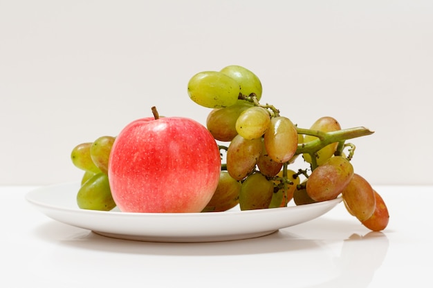 Cacho de uvas verdes e uma maçã em um prato com o fundo branco.