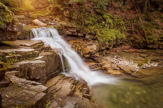 Cachoeira da montanha em cascata com raios de sol passando pelas folhas da manhã. A água está borrada devido à longa exposição.