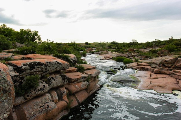 Cachoeira flui entre rochas altas, pedras vermelhas e penhascos, arbustos verdes e árvores