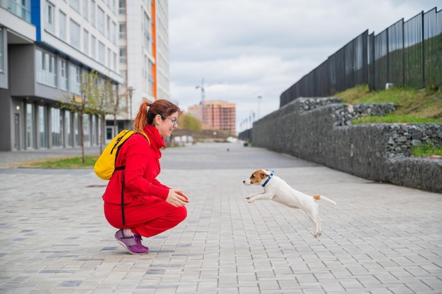 Cachorro inteligente Jack Russell Terrier juega con el dueño en la calle Un perro de pelo corto de pura raza salta a los brazos de una mujer europea en un chándal rojo En movimiento