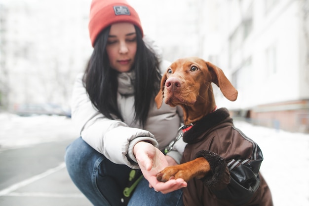 Cachorro marrom com um vestido e uma garota feliz sentada na neve no inverno e brincando