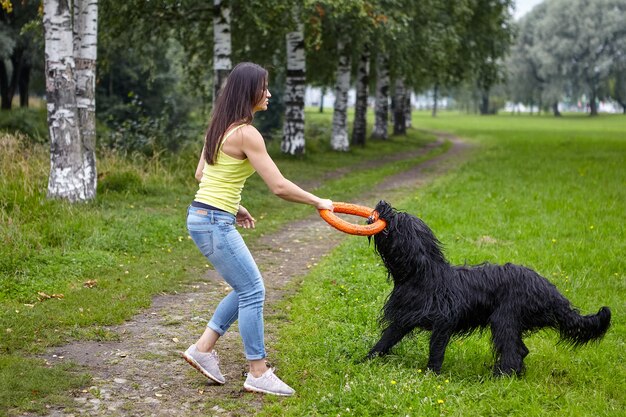 Cachorro peludo preto tem treinamento com jovem branca com ajuda de brinquedo durante uma caminhada ao ar livre.