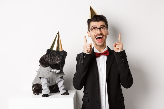 Cachorro pug preto bonito usando cone de festa e em pé perto do feliz proprietário, homem apontando os dedos para o espaço da cópia, comemorando o aniversário, fundo branco.