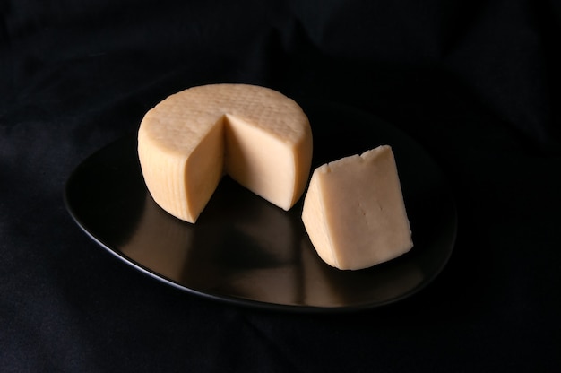 Caciotta-Käse auf einem schwarzen Teller. Vom Käse wird ein dreieckiges Stück abgeschnitten. Schwarzer Hintergrund. Isoliert. Nahaufnahme.