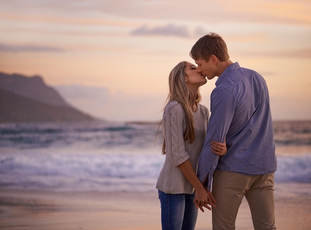 Cada beso se siente como nuestra primera toma de una pareja joven disfrutando de un beso romántico en la playa al atardecer.