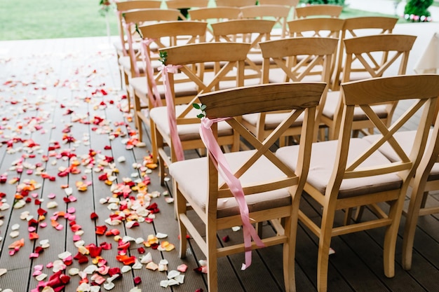 Cadeiras de madeira são decoradas com fitas de laço rosa e verdes na área de banquete do quintal Cerimônia de casamento Pétalas de rosas espalhadas na estrada decoração de casamento Vista lateral e vista traseira