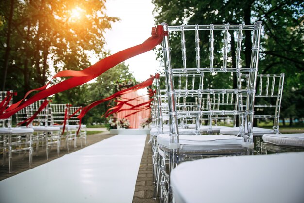 Cadeiras elegantes transparentes decoradas com fitas vermelhas antes da cerimônia de casamento ao ar livre