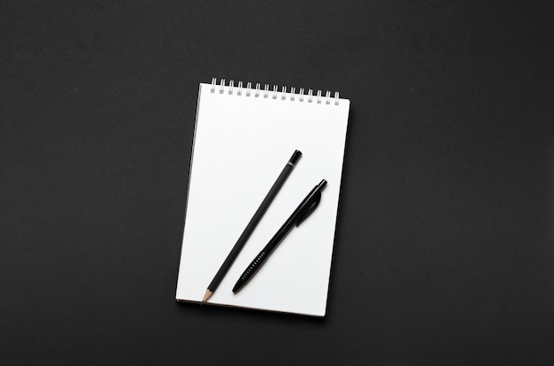 Caderno aberto branco com lápis preto e caneta em fundo preto. Mesa de escritório, artigos de papelaria. Vista superior, plana leigos.