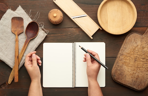 Caderno aberto com folhas brancas em branco e utensílios de cozinha em uma mesa de madeira marrom, mãos femininas segurando um lápis preto, vista superior