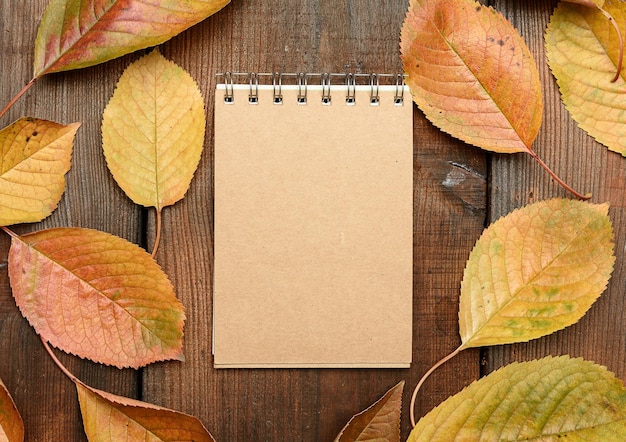 Foto caderno aberto com folhas marrons em branco e folhas de cereja amarelas em fundo de madeira marrom vista superior