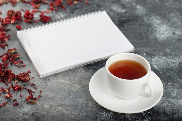 Caderno aberto com uma xícara de chá e pétalas de flores colocadas na mesa de pedra.