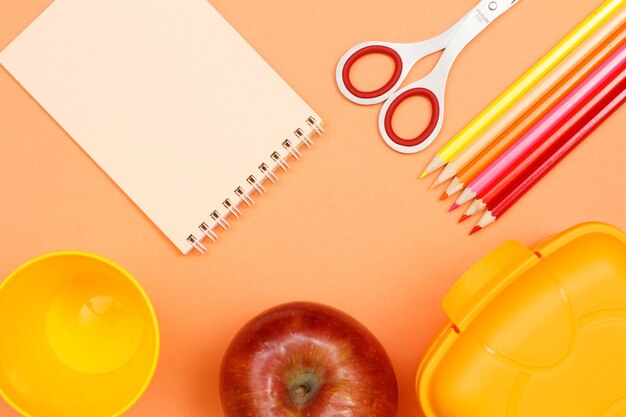 Caderno, apple, tesoura, lápis de cor, copo plástico e lancheira em fundo rosa.