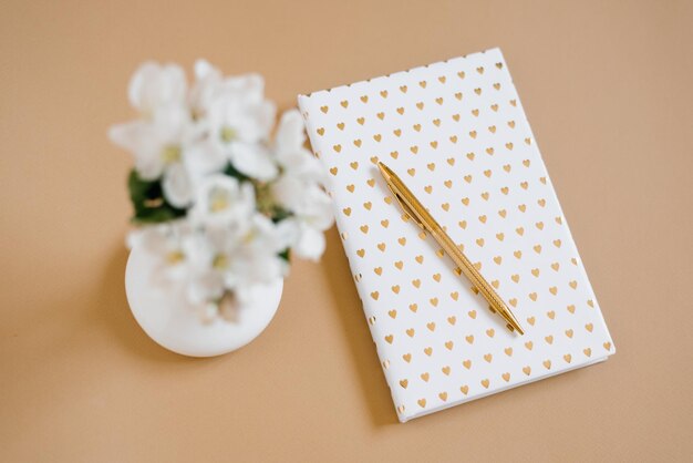 Caderno branco com corações de ouro na capa, caneta de ouro e flores de maçã branca em um vaso fora de foco em uma mesa bege. Conceito de layout e design. Postura plana.