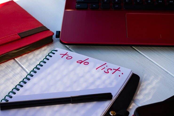 Foto caderno com a lista de tarefas pendentes com telefone, diário vermelho e laptop em um fundo branco de madeira.