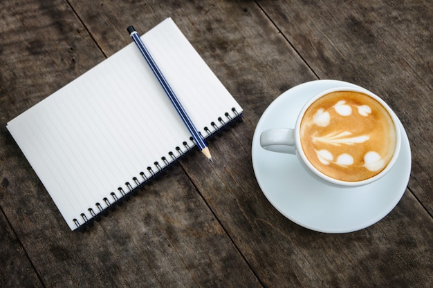 Foto caderno com lápis e xícara de café de padrão de coração na mesa de madeira velha.