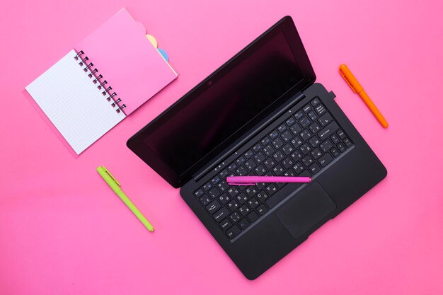 Caderno de laptop e canetas em um fundo rosa