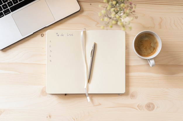 Caderno de papel em branco de vista superior com lista de tarefas, flores, teclado de laptop, xícara de café e caneta.