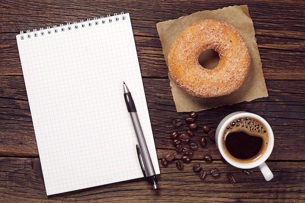 Caderno e xícara de café quente com donut na velha mesa de madeira, vista superior