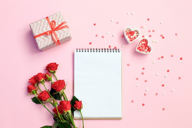 Caderno em branco aberto com caixa de presente, flores e corações em um fundo rosa