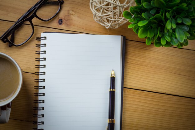 Caderno em branco com caneta e óculos ao lado da xícara de café na mesa de madeira