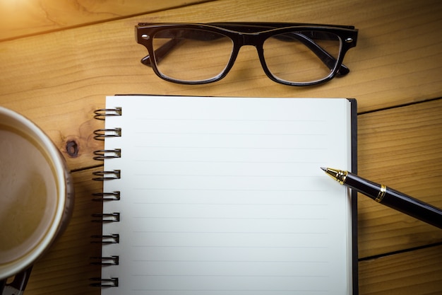 Caderno em branco com caneta e óculos ao lado da xícara de café na mesa de madeira