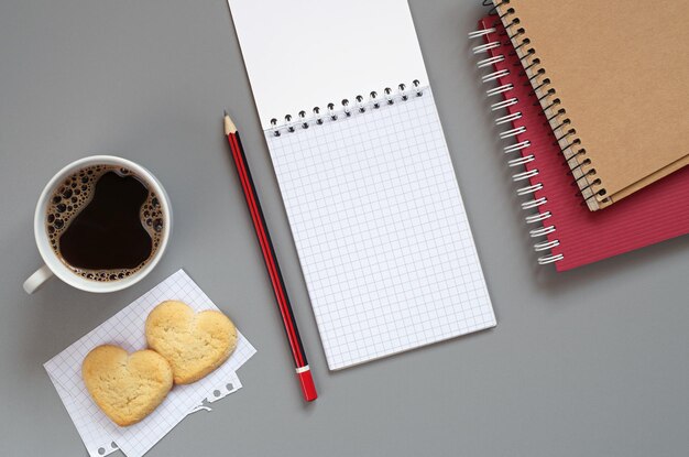 Caderno, lápis e xícara de café com biscoitos em forma de coração na mesa cinza, vista superior