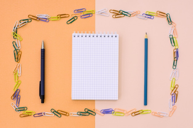 Foto caderno na gaiola com caneta e lápis em um quadro de clipes de papel coloridos em um fundo duplo bege e laranja conceito de volta à escola