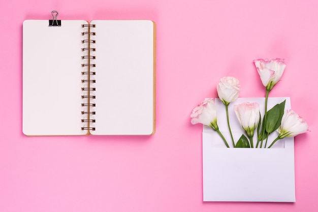 Caderno vazio aberto e buquê de flores eustoma em envelope na superfície rosa.