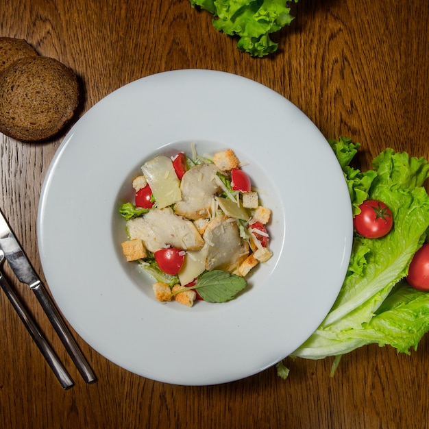 Caesar-Salat mit Hähnchenfilet auf weißem Teller mit Salat und Brot daneben und Silberbesteck