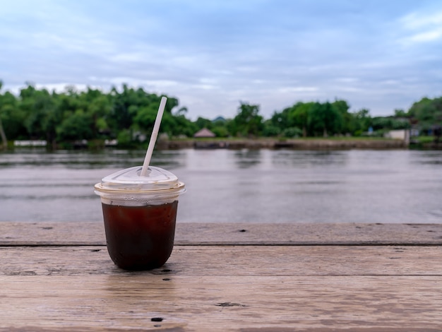 Café americano helado en taza transparente para llevar en la mesa de madera con vistas frescas al río tranquilo