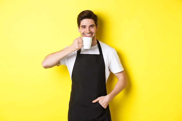 Café barista bebiendo una taza de café y sonriendo, vistiendo delantal negro, de pie sobre la pared amarilla