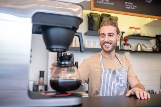 Café, café. homem jovem adulto sorridente com avental com cafeteira perto da cafeteira atrás do balcão de um café