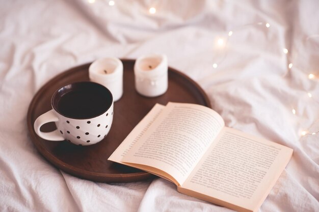 Café caliente con libro abierto y velas sobre brillantes luces de Navidad de cerca. Hora del desayuno.