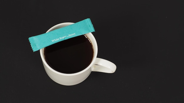 Café caliente en taza blanca y bolsita de azúcar blanca sobre fondo negro.